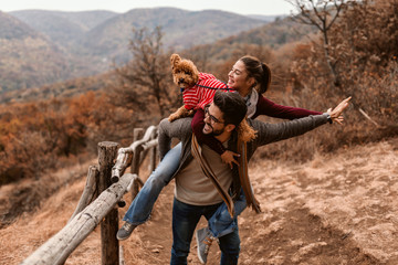 Couple having piggyback in nature. On man's backs woman and poodle. In background forest and mountain. Autumn time.