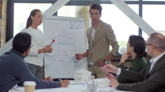 Young male and female architects explaining building construction plan on flipchart and discussing it with team of professionals while giving presentation in the office