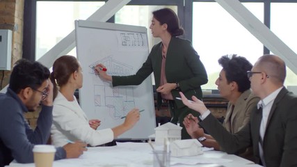 Young female architect drawing building construction plan on flipchart and discussing it with diverse team of colleagues while giving presentation in the office