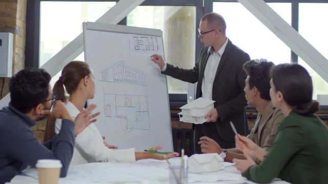 Caucasian male architect in eyeglasses and formal suit standing by flipchart and discussing house model with interracial team of colleagues while giving presentation during office meeting