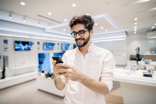 Smiling man standing in tech store with smart phone in his hands. New technologies concept.