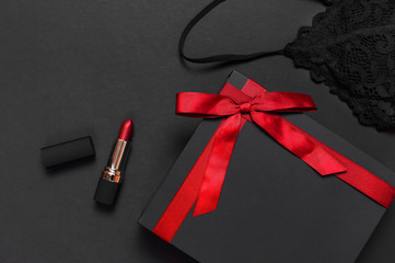 Black gift box with red ribbon, lace bra underwear, red lipstick on dark background top view flat lay with copy space. Female essential erotic accessories, fashionable underwear, gift to woman.