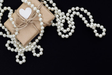 Gift with white heart in craft paper and pearl necklace on the black background. Romance, love concept. Top view, flat lay, copy space, card layout