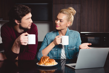 attractive girl holding coffee cup, pointing at laptop screen and looking at husband in kitchen