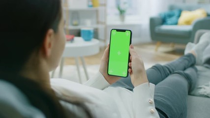 Young Woman at Home Resting on a Couch using with Green Mock-up Screen Smartphone. Girl Using Chroma Key Mobile Phone, Internet Browsing, Posting on Social Networks. Point of View Camera Shot. - Powered by Adobe