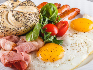 Breakfast, fried eggs, bun, sausage, bacon, prosciutto, fresh salad on plate on grey table surface. Healthy food, top view, flat lay
