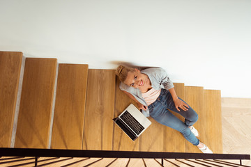 overhead view of attractive girl sitting with laptop on stairs and looking at camera