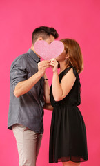 Young couple hiding behind pink heart on color background. Celebration of Saint Valentine's Day