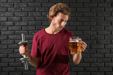 Young man with dumbbell and mug of beer near black brick wall. Concept of choice between alcohol...