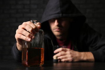Depressed young man drinking whiskey at table near black brick wall. Alcoholism concept