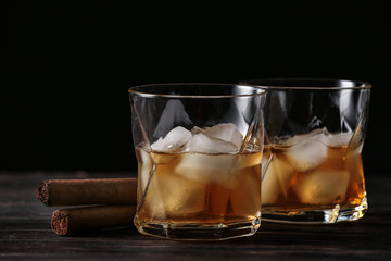 Glasses of cold whiskey with cigars on wooden table against dark background