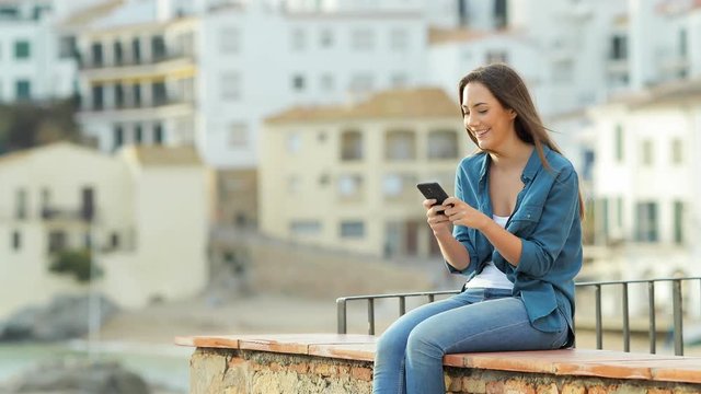 Happy woman texting on smart phone outdoors sitting on a ledge in a coast town