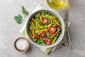 Salad with asparagus, bell pepper, cherry tomatoes, red onion and green peas in ceramic bowl on stone background. Top view. Copy space.