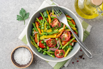 Salad with asparagus, bell pepper, cherry tomatoes, red onion and green peas in ceramic bowl on stone background. Top view. Copy space.