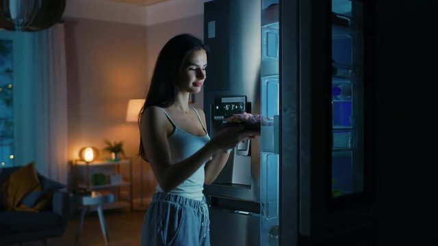 Beautiful Young Woman Comes to the Kitchen in the Evening. She is Hungry and Opens the Fridge. Takes out a Piece of a Delicious Creamy Cake and Bites It. She Feels Satisfied.