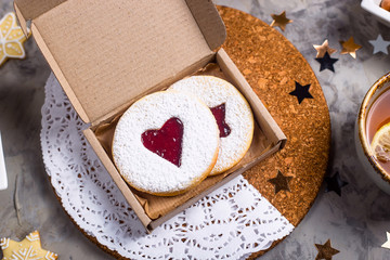 Round cookies with heart-shaped jam in a gift box among Christmas decorations, candles and cups of tea on a gray table. Homemade baking concept. Top view, flat lay