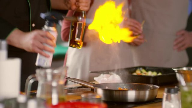 Medium shot of male chef pouring alcohol on frying vegetables, setting fire on dish, then taking pan in hand and shaking it while seniors on background are watching