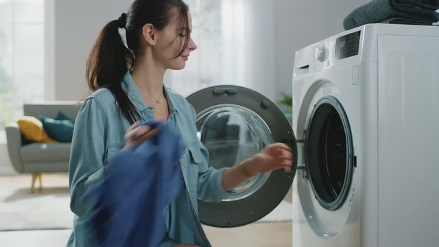 Close Up Shot of a Beautiful Smiling Brunette Woman Sitting in Front of a Washing Machine in Homely Jeans Clothes. She Loads the Washer with Dirty Laundry. Bright Living Room with Modern Interior.