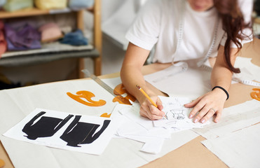 Female designer drawing pencil sketches of clothing sitting at big table with flat paper patterns, measuring ruler and curves on blurred background of tailor design studio interior.