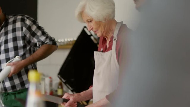 Medium shot of aged woman in apron standing at table, cutting zucchini, then male chef with dripping-pan in his hands showing her next steps of cooking