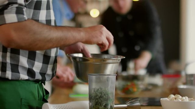 Tilt up of Arab man putting with his hands cut pieces of greens in special plastic jar to chap them with immersion blender, while cooks on background talking
