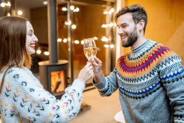 Young couple in sweaters celebrating winter holidays standing with sparkling wine in the modern house with fire place