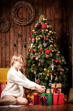 Beautiful pregnant woman wearing a warm sweater and socks, sitting near tha Christmas tree holding gift boxes