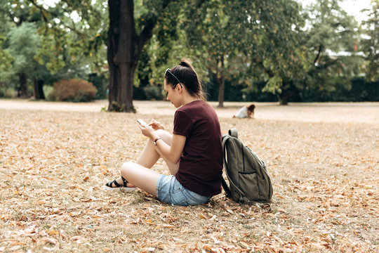 A girl with a backpack sits in an autumn park and uses a cell phone to make a call or be online.