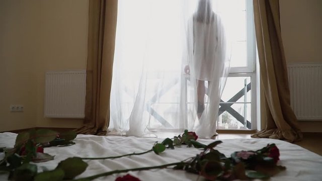 Pretty young woman in short transparent shirt standing near a large bright window. Girl in nightie looks out the window while standing in the bedroom in the morning.