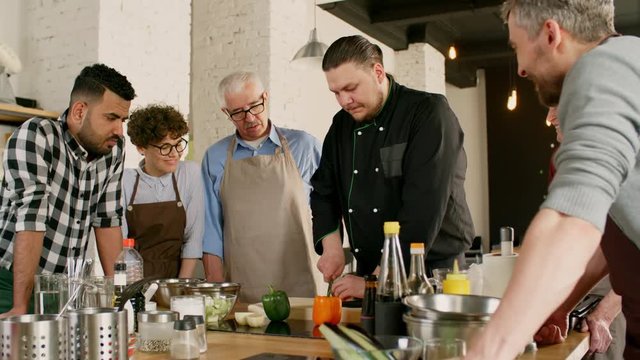 Medium shot of people of different ages and nations gathered around male chef who is showing how to slice something correctly