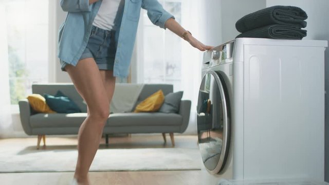 Beautiful Young Woman Sits on Her Knees Next to the Washing Machine. She Loaded the Washer with Dirty Laundry and Configured the Wash With Her Smartphone. Shot in Living Room with Modern Interior.