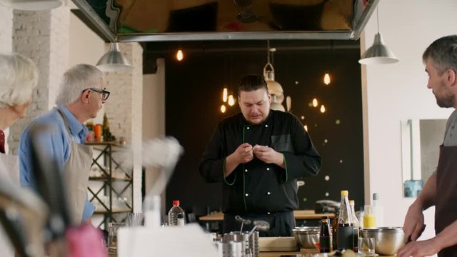 Medium shot of people of different ages gathered around kitchen table and watching chef showing how easily peel the garlic