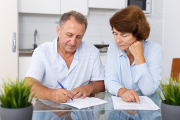 Fototapeta na wymiar Smiling mature couple at table in home kitchen filling up documents