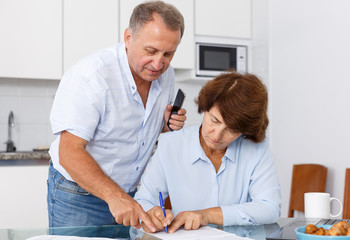 Fototapeta premium Mature couple at table in home kitchen, woman filling up documents