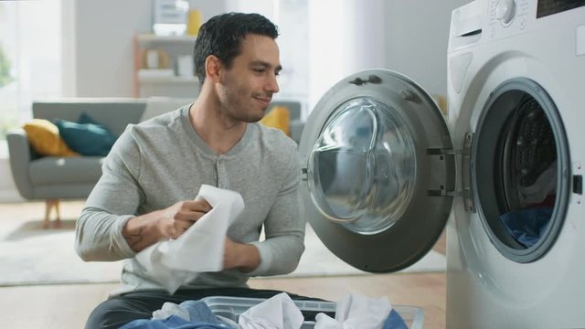 Handsome Smiling Young Man in Grey Jeans and Shirt Sits in Front of a Washing Machine at Home. He Loads the Washer with Dirty Laundry. Bright and Spacious Living Room with Modern Interior.