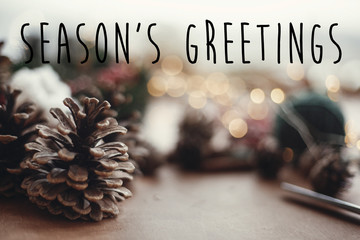 Season's greetings text sign on pine cones on background of christmas wreath,lights, fir branches,...