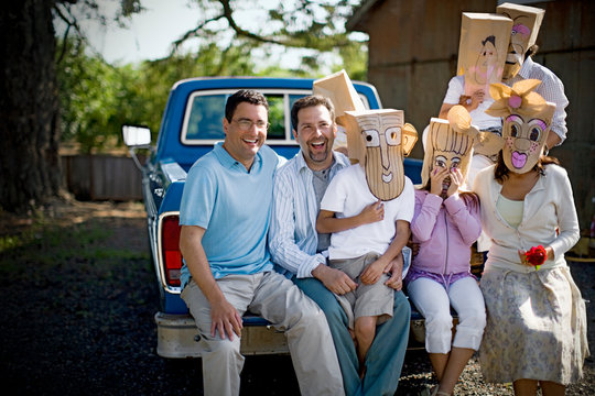 Portrait of two smiling mid-adult men sitting with their family who are wearing paper bag masks on the back of a truck.