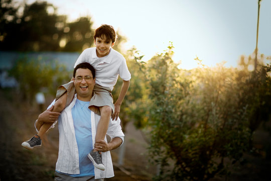 Portrait of a smiling mid-adult man holding his son on his shoulders while standing in an apple orchard.