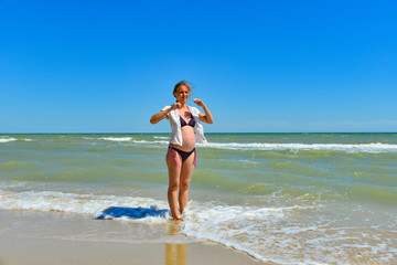 Fototapeta premium A happy and joyful pregnant woman stands by the sea and ocean on the beach under the sun in the open air, hair develops in the wind.