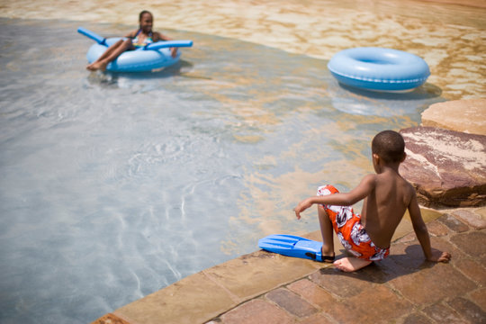 Young boy sitting at the edge of a swimming pool wearing a single flipper.