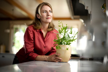 Woman holding a pot plant on a kitchen counter and looking out of the window.