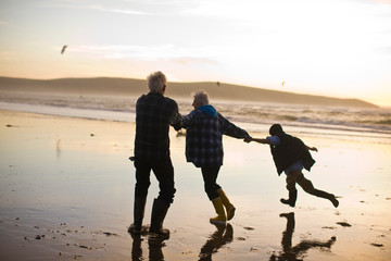 Grandparents having fun while holding hands with their grandson on a beach at sunset.