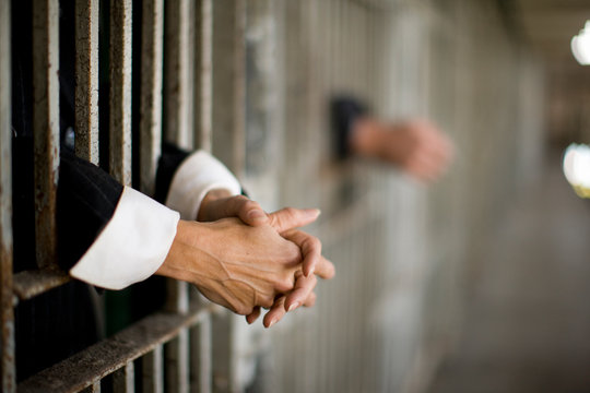 Hands of a mid-adult woman through bars of a prison cell in a derelict building.