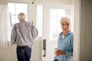 Portrait of a senior woman drinking coffee as her husband looks out a window.
