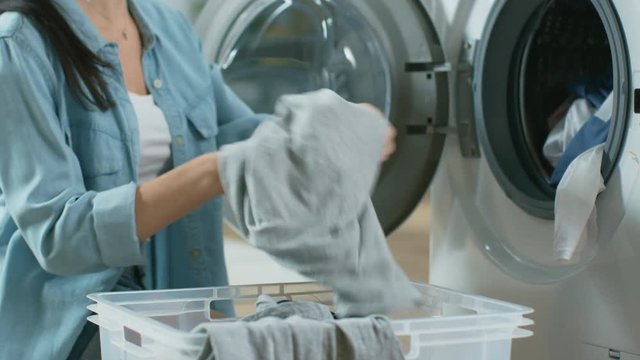 Close Up of a Beautiful Young Woman in Homely Clothes Sitting in Front of a Washing Machine. She Loads the Washer with Dirty Laundry. Bright and Spacious Living Room with Modern Interior.