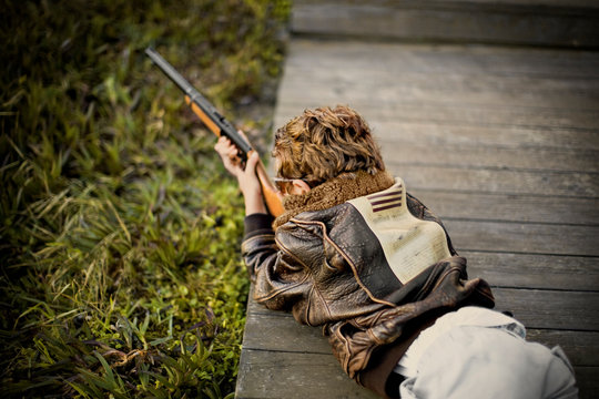 Boy aiming a shotgun while lying on his stomach on top of a wooden footbridge.