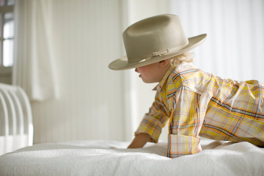 Young boy crawling along a bed wearing a large hat.