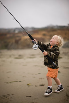 Happy young toddler holding a fishing rod while standing on a remote sandy beach.