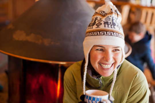 Laughing mid adult woman holding a mug of coffee while wearing a knitted hat indoors.