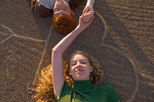 Portrait of a teenage couple holding hands lying on their backs on a heart shape drawn in the dirt.